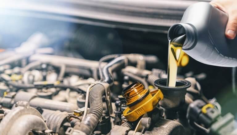 A person pours engine oil from a gray bottle into a car engine. The engine compartment is visible with various components and hoses. Natural light illuminates the scene, highlighting the oil's golden color as it flows into the engine.