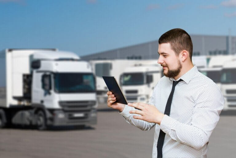 A bearded man in a white shirt holds a tablet and smiles, standing in front of a row of white trucks parked at a logistic center on a clear day.