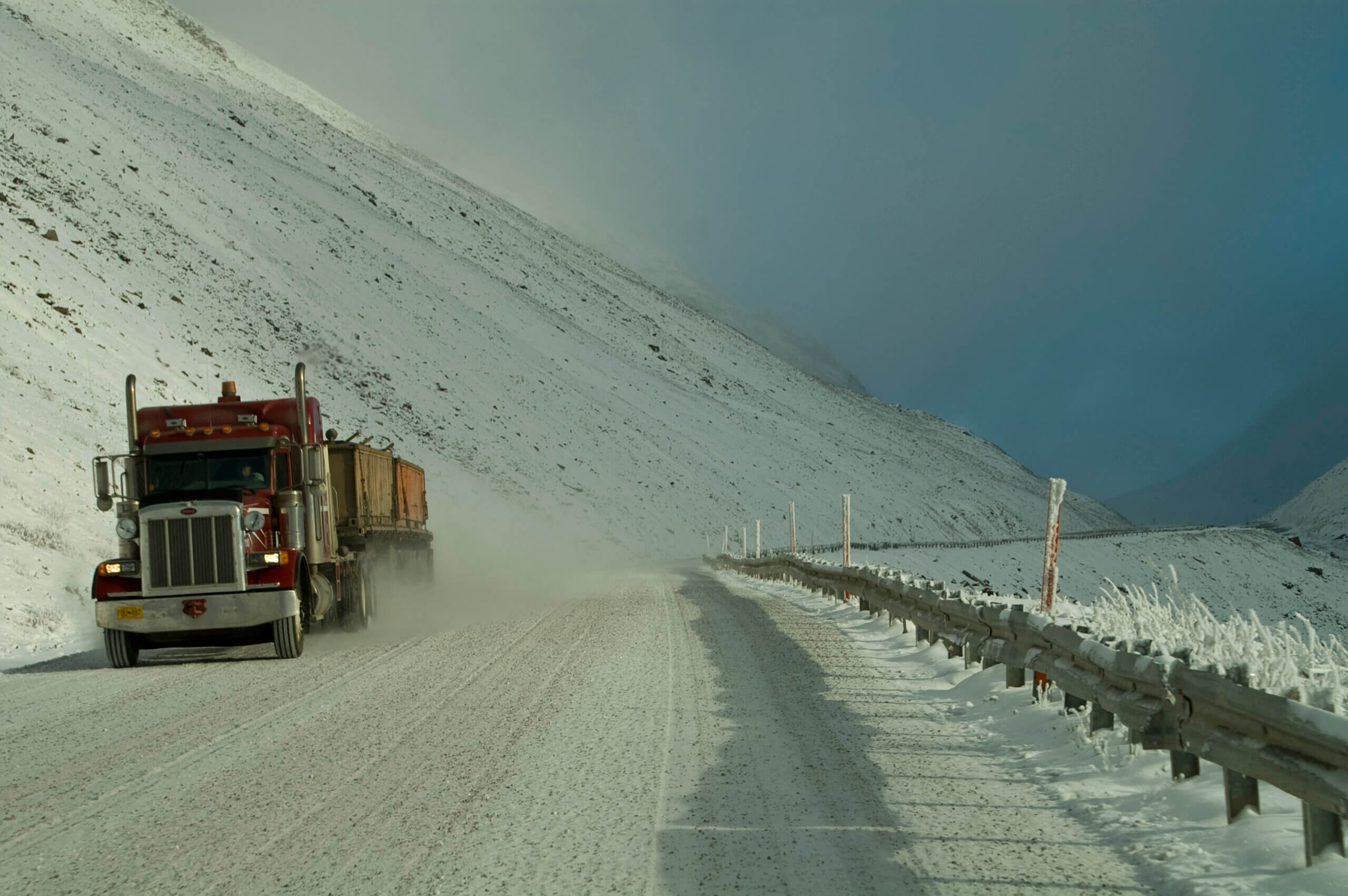 A red semi-truck drives through a snowy, mountainous landscape on a wintery day. The road is covered in snow, and the sky is overcast, with a hint of blue visible in the distance. The scene conveys a sense of cold and isolation.