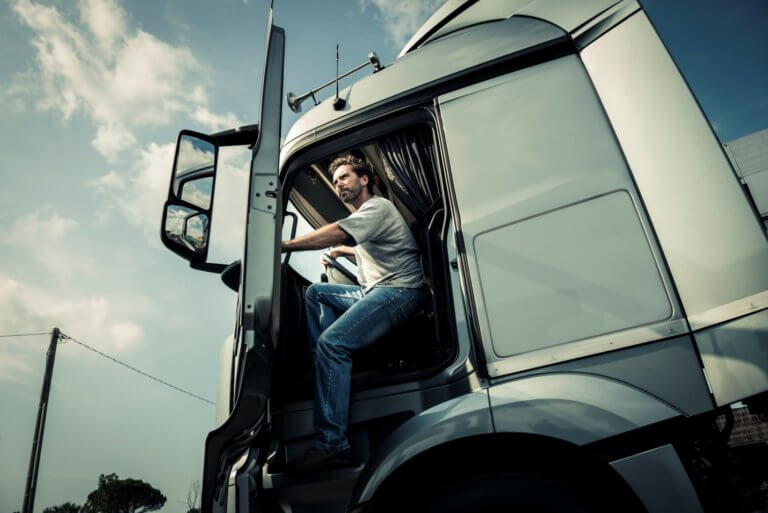 A man sits in the driver’s seat of a large truck, looking out of the open door. The sky is partly cloudy, and the truck is parked on a street with utility poles in the background. The scene is set during the day.