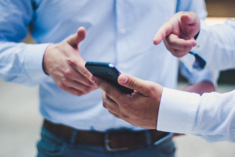 Two people in blue shirts are standing close to each other. One person is holding a smartphone while the other points towards it, indicating a discussion or explanation. The focus is on their hands and the phone.