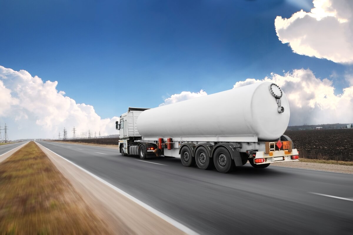 A large white tanker truck drives down an empty highway under a blue sky with scattered clouds. The road stretches into the distance alongside an open field.