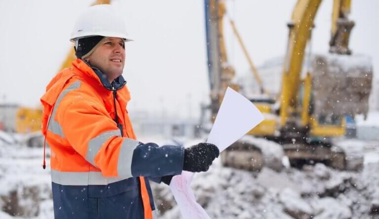 A construction worker in an orange safety jacket and white helmet stands in a snowy environment, holding a set of papers. Excavators are blurred in the background, and snowflakes are falling around him.