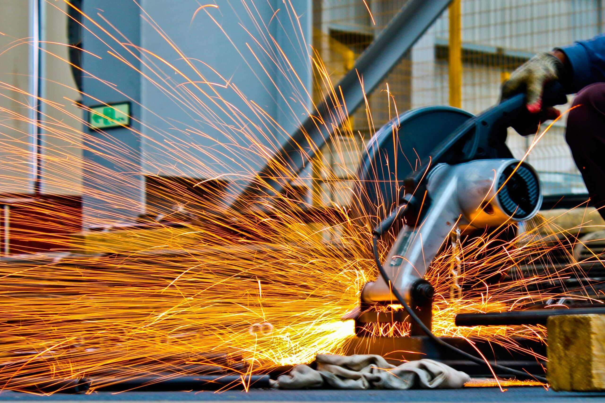 A person uses a power tool to cut through metal, creating a shower of orange sparks. The background features industrial elements with a cage-like structure, and safety equipment is visible.