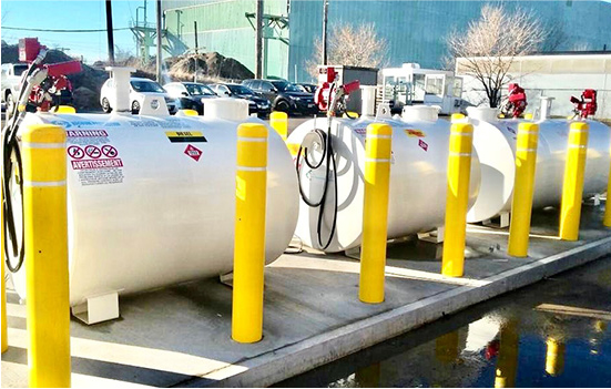 A row of three large white fuel storage tanks with attached dispensers is lined up outdoors. Yellow safety bollards are positioned in front of each tank. Vehicles and a building are visible in the background.