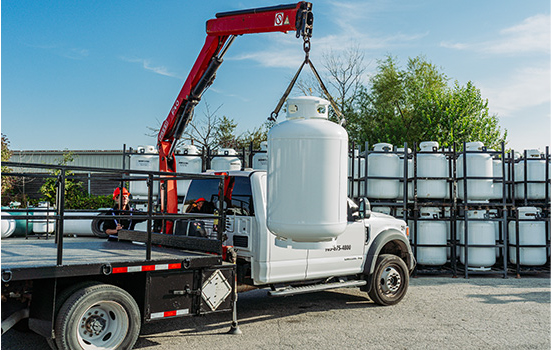 A flatbed truck with a red crane is lifting a large white propane tank. In the background, multiple propane tanks are stacked on metal racks. The scene is outdoors, with a clear sky and some trees visible.