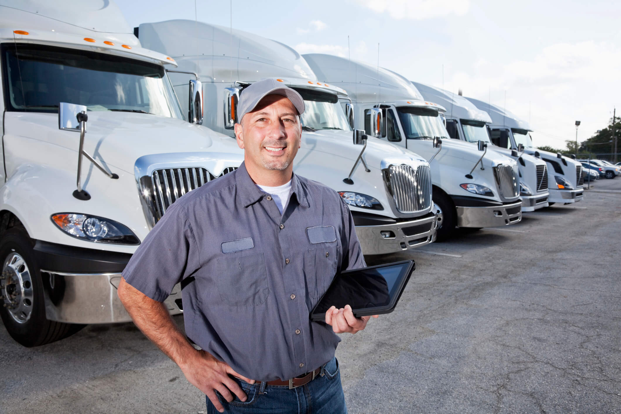 A truck driver is standing in a large parking lot in front of several large trucks. He is facing the camera, and he is wearing a short-sleeved blue shirt, blue jeans, a brown belt and a gray hat. He is holding a black tablet with his left hand. There are six big rigs seen lined up behind him, going from left to right. The large trucks all look the same, being all white and having silver grills and mirrors. There are multiple cars seen parked to the right of the trucks. A cloudy blue sky is seen in the background.